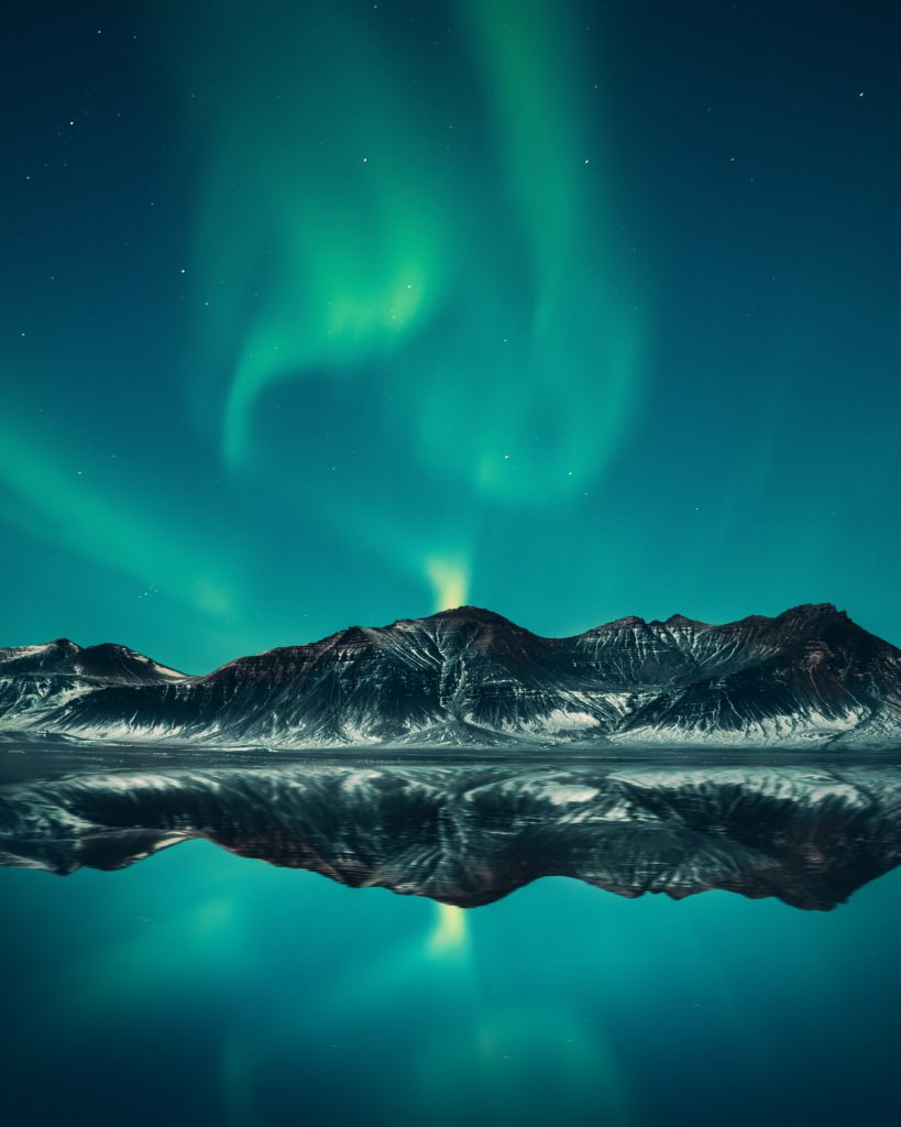 A blue-green northern light connecting to a mountain by a lake.